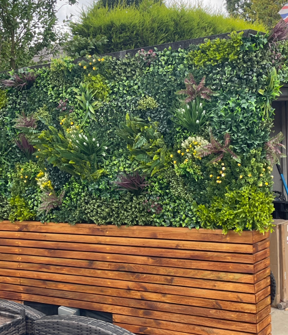 Artificial green wall panel outdoors with dense mixed leaves and yellow flower clusters