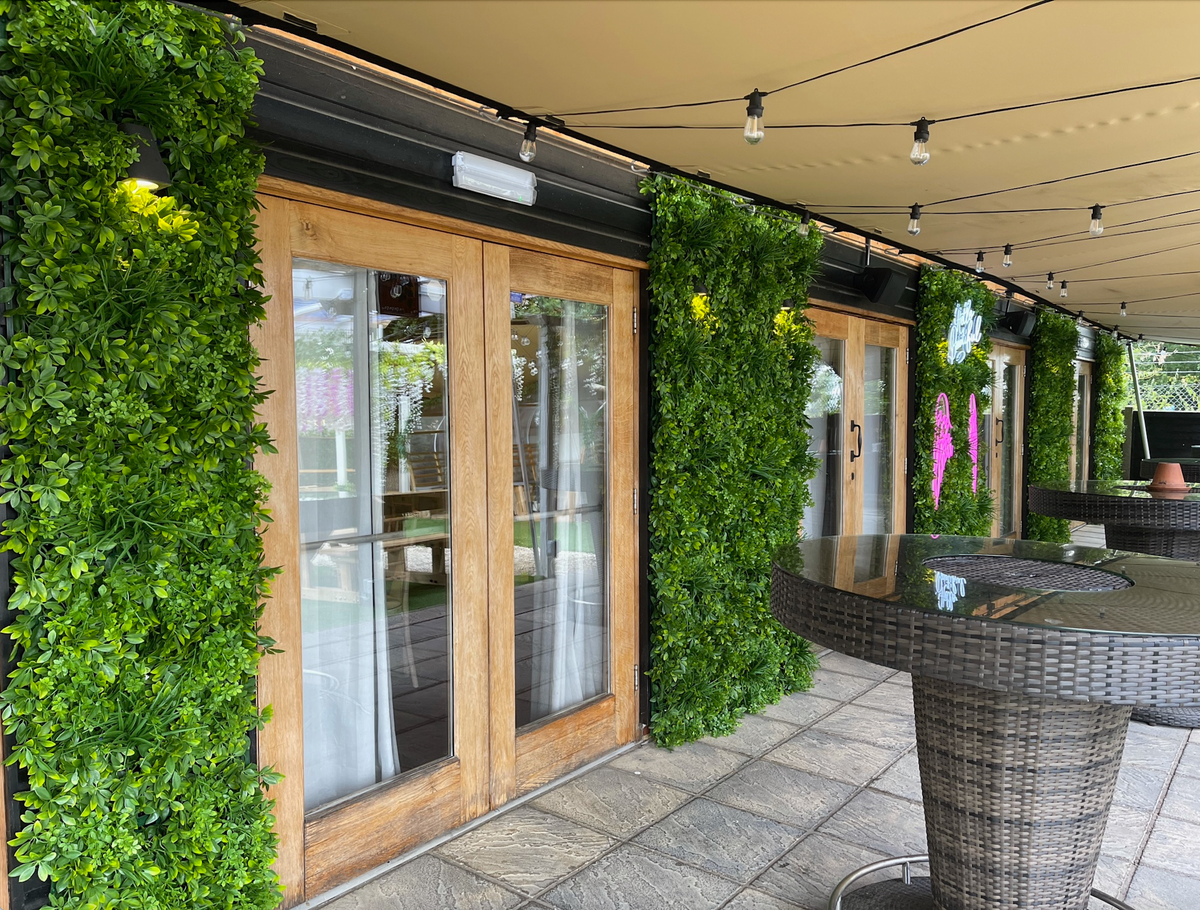 Artificial green foliage wall panels forming a vertical strip along windows, dense 3D leaves