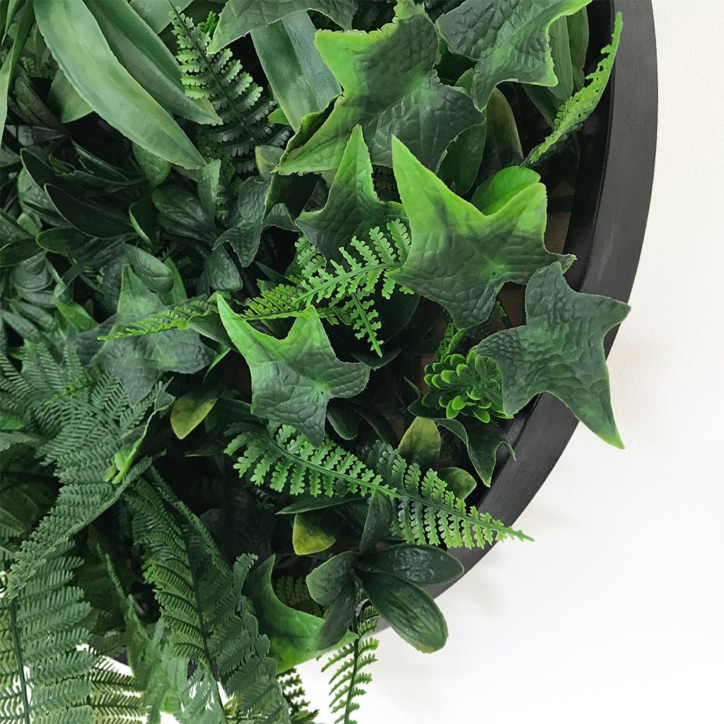 Black round wall art panel with dense green artificial leaves and ferns