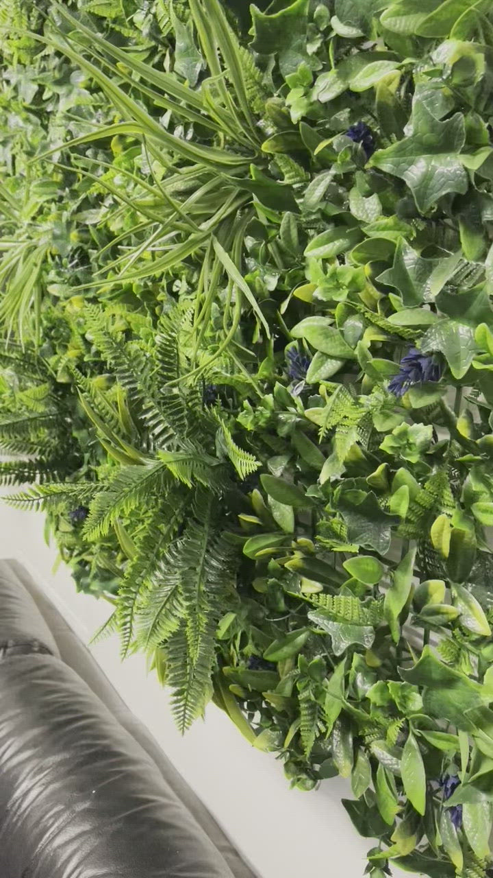 Artificial green wall panel with variegated greens of ivy, ferns, palm heads, grasses & blue-purple flowers  100x100 cm