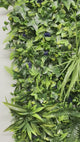 Artificial green wall panel with variegated greens of ivy, ferns, palm heads, grasses & blue-purple flowers  100x100 cm