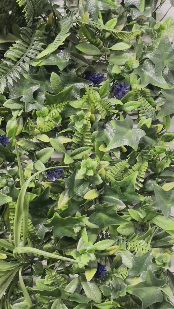 Artificial green wall panel with variegated greens of ivy, ferns, palm heads, grasses & blue-purple flowers  100x100 cm